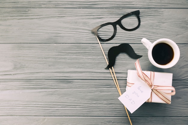 Photo gratuite lunettes décoratives et moustache près du présent avec une tasse de boisson