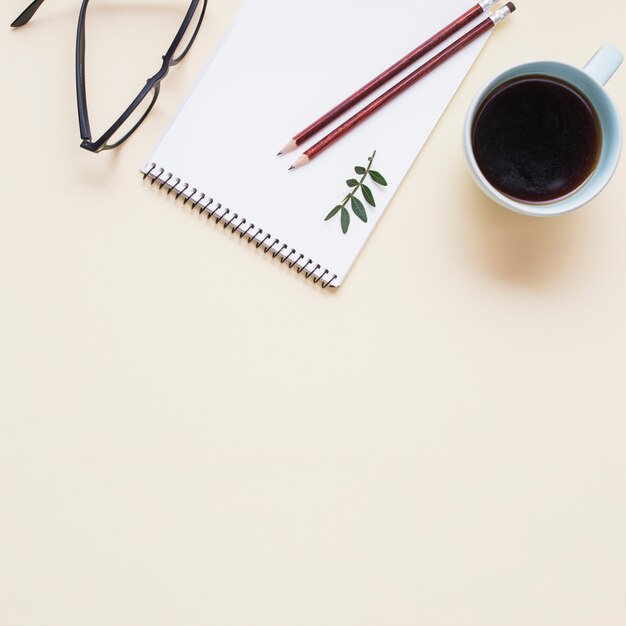 Lunettes noires; tasse à thé et deux crayons sur le bloc-notes en spirale sur fond beige