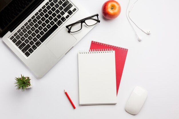 Lunettes de vue sur ordinateur portable, pomme, écouteurs, crayon de couleur, bloc-notes à spirale et souris sur un bureau blanc