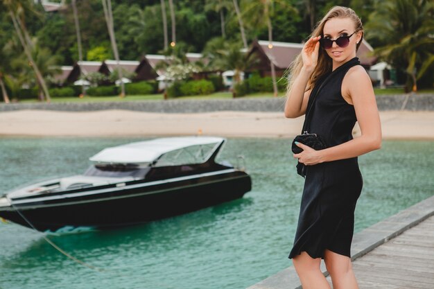 Luxe sexy jolie femme vêtue d'une robe noire posant sur la jetée dans un hôtel de luxe, lunettes de soleil, vacances d'été, plage tropicale