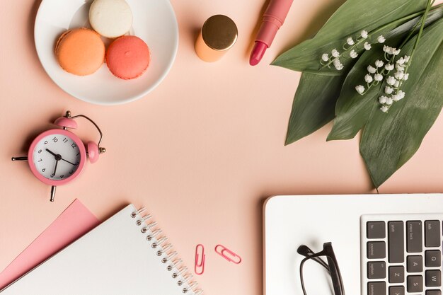 Macarons, réveil, rouge à lèvres, feuilles et fleurs avec bloc-notes en spirale et ordinateur portable