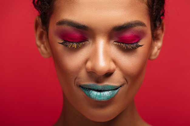 Macro charmante femme afro-américaine étant à la mode et à la mode portant des cosmétiques colorés posant avec les yeux fermés, sur le mur rouge