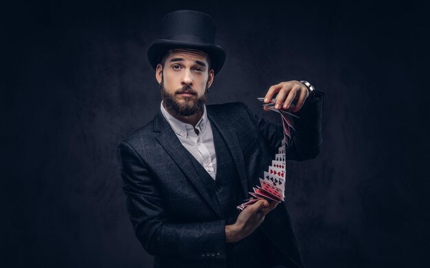 Un magicien barbu en costume noir et chapeau haut de forme, montrant un tour avec des cartes à jouer sur fond sombre.