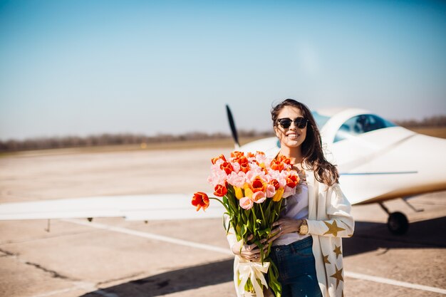 Magnifique brune se dresse avec un grand bouquet de tulipes devant un avion