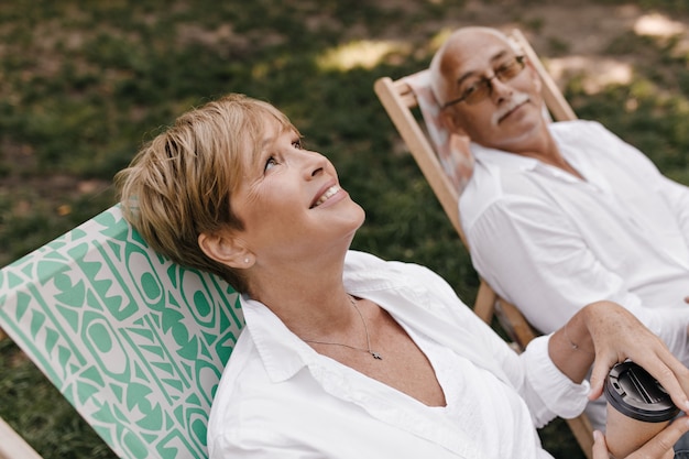 Photo gratuite magnifique femme aux cheveux blonds en vêtements blancs souriant, tenant une tasse de thé et assis sur une chaise avec un homme aux cheveux gris avec des lunettes en plein air.