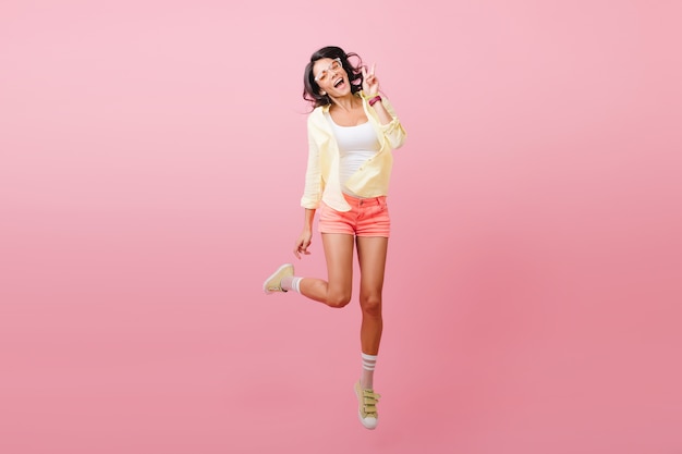 Magnifique femme aux cheveux noirs en tenue tendance debout sur une jambe pour la photo. Portrait intérieur de joyeuse jeune femme latine dans des chaussures jaunes élégantes.