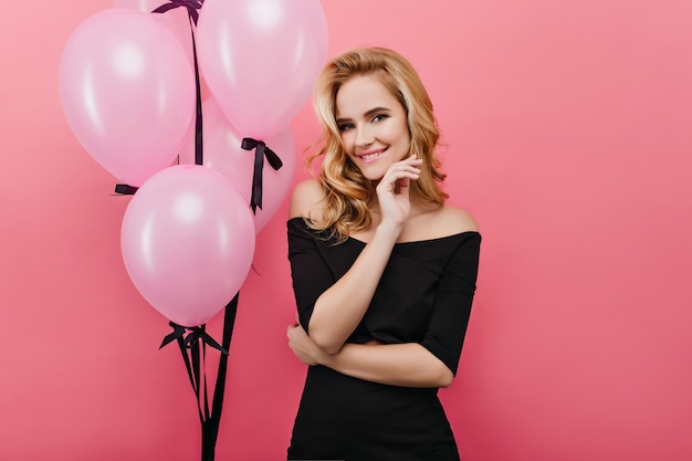 Magnifique femme blonde aux cheveux bouclés debout sur un mur lumineux pendant la fête d'anniversaire. Superbe femme blonde célébrant les vacances avec des ballons.