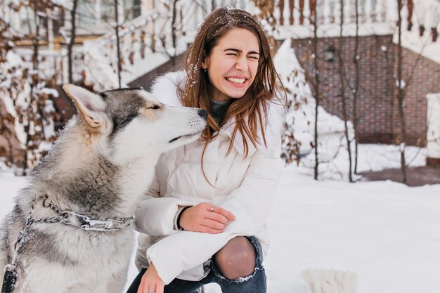 Magnifique femme en blouse blanche bénéficiant d'une promenade hivernale avec son drôle de chien. Portrait en plein air de belle femme européenne jouant avec husky à cour enneigée.