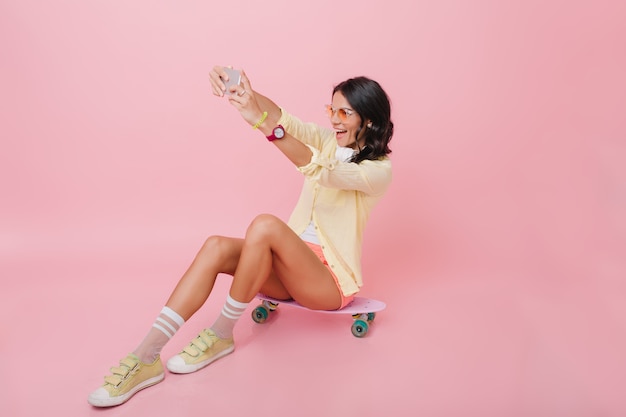 Magnifique Femme Brune En Tenue Jaune Assis Sur Une Planche à Roulettes Dans La Chambre Avec Intérieur Rose. Portrait Intérieur D'une Fille Rêveuse En Chaussettes Mignonnes Faisant Selfie.