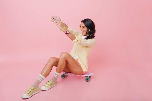 Magnifique femme brune en tenue jaune assis sur une planche à roulettes dans la chambre avec intérieur rose. Portrait intérieur d'une fille rêveuse en chaussettes mignonnes faisant selfie.