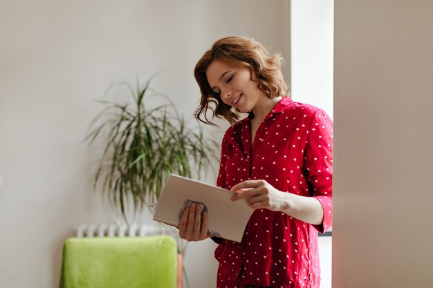 Magnifique femme en pyjama lisant le magazine avec intérêt. Plan intérieur d'une adorable femme bouclée à la maison.