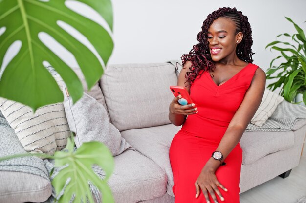 Magnifique jeune femme africaine en robe rouge luxueuse dans un appartement de luxe assis sur un canapé avec téléphone portable Mode beauté