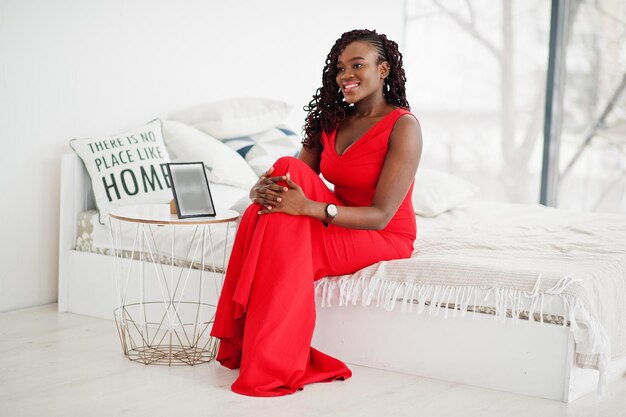 Magnifique jeune femme africaine en robe rouge luxueuse dans un appartement de luxe assis sur le lit Mode beauté