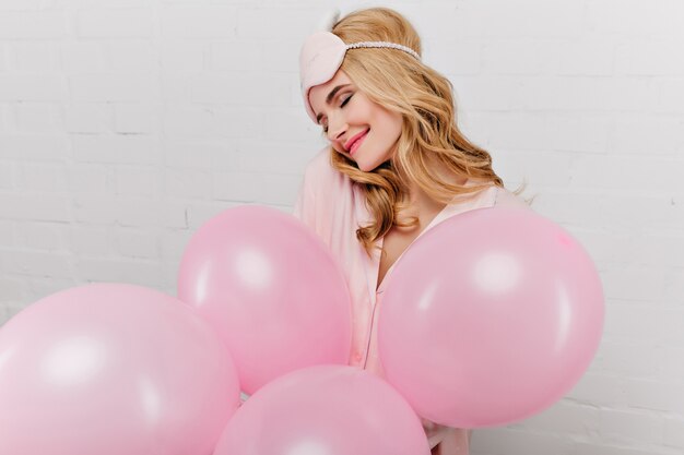 Magnifique jeune femme en masque rose posant avec plaisir, tenant des ballons d'hélium. Portrait intérieur d'une superbe fille célébrant son anniversaire le matin.