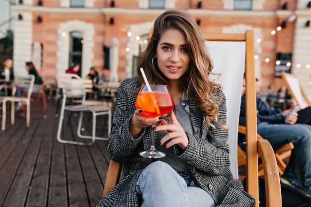 Magnifique modèle féminin aux yeux bleus en manteau gris assis dans un café en plein air confortable