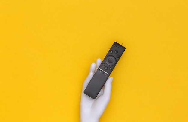 La main du mannequin blanc tient la télécommande du téléviseur sur fond jaune. vue de dessus