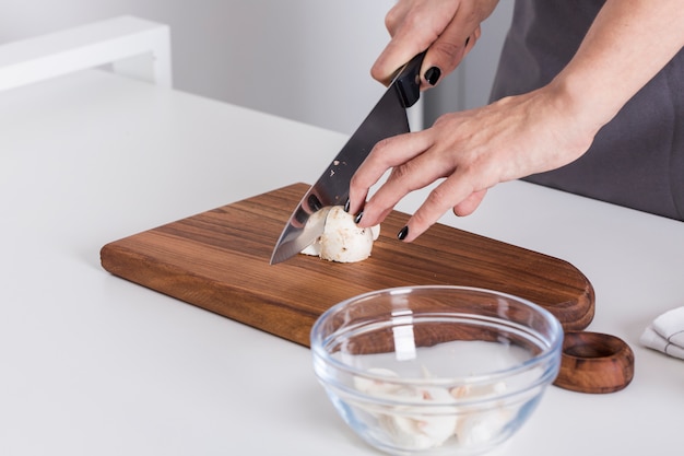 Main de femme coupant le champignon avec un couteau sur une planche à découper sur la table blanche