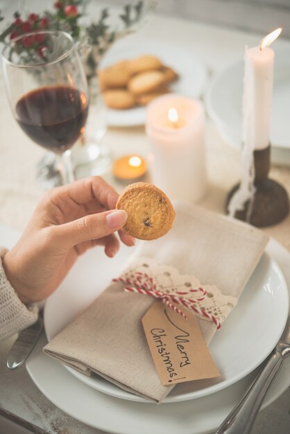 Main de femme non reconnaissable tenant un biscuit contre une table dressée pour le dîner de Noël