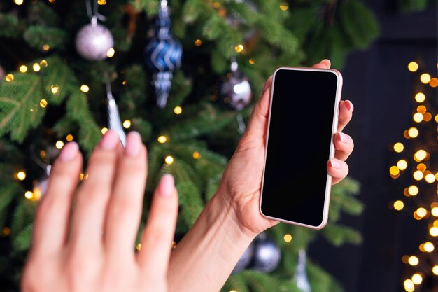 La main d'une femme salue l'écran du smartphone sur fond d'arbre de noël. le concept de salutations en ligne. coronavirus et quarantaine pour la nouvelle année. espace vide pour la conception
