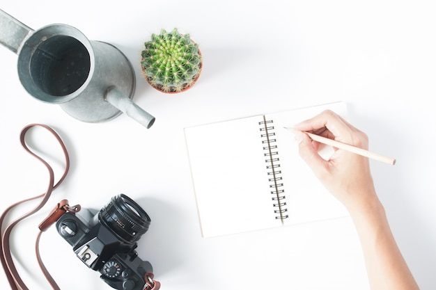 La main de la femme tenant un crayon sur un cahier vide avec une caméra de film, un pot de cactus et un rivage vintage, isolé sur fond blanc, vue de dessus, plat