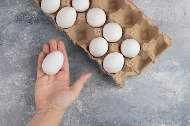 Main de femme tenant un œuf de poule blanc frais sur un marbre.