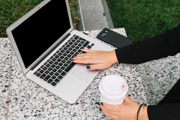 Main de femme tenant une tasse de café jetable en tapant sur un ordinateur portable