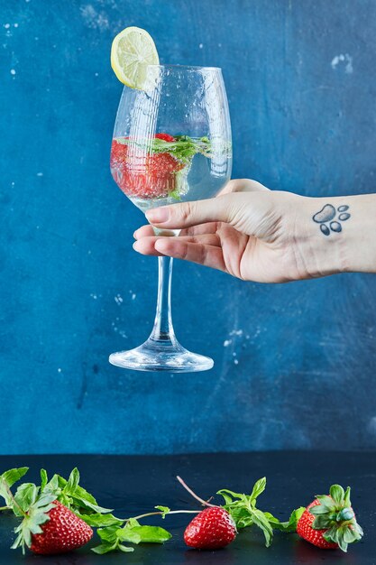 Main de femme tenant un verre de jus avec des fruits entiers et de la menthe à l'intérieur