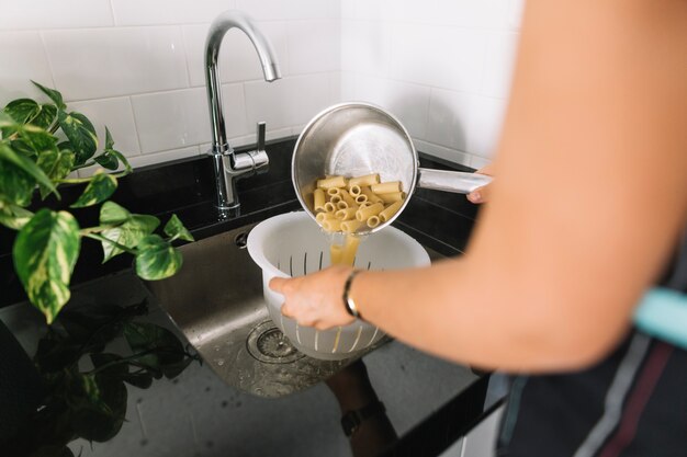 Main de femme versant des pâtes rigatoni dans la passoire blanche de la casserole