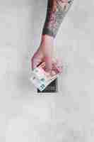 Photo gratuite main de l'homme avec tatouage tenant des billets d'euro et passeport sur le mur de béton
