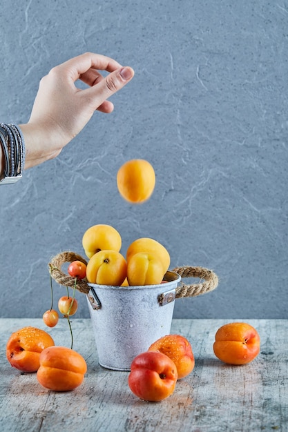 Photo gratuite main jetant des abricots dans le petit seau avec des nectarines sur une surface en marbre