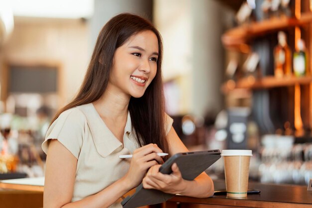 Main de nomade numérique pigiste asiatique à l'aide d'une tablette travaillant à distance n'importe où workation s'asseoir au comptoir près d'un bar à café contacter un client à l'étranger avec un accord de réussite paisible et souriant
