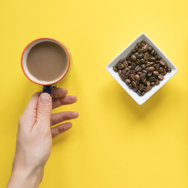 Main de la personne prenant une tasse de café avec des grains de café torréfiés sur fond jaune