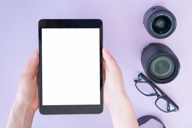 Photo gratuite main de la personne tenant une tablette numérique sur la lentille de la caméra et des lunettes sur fond lavande