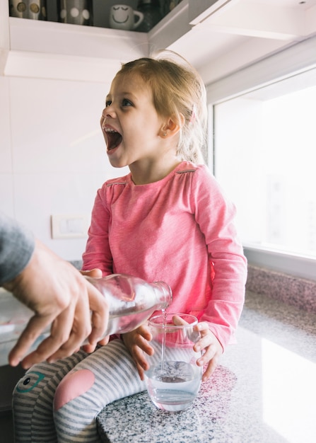 Main d&#39;une personne versant de l&#39;eau dans le verre alors que la fille assise sur le comptoir de la cuisine