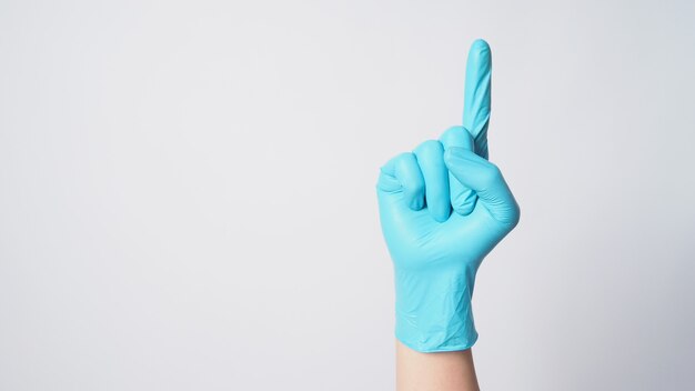 La main porte un gant chirurgical bleu et fait un signe de main avec un doigt ou pointe vers le haut sur fond blanc.
