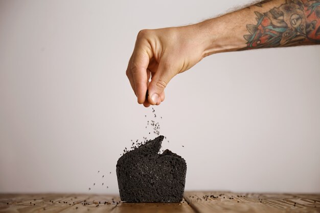 Main tatouée verse des épices de graines noires sur le dessus du pain fait maison biologique de charbon noir isolé sur du papier craft sur table en bois en boulangerie artisanale