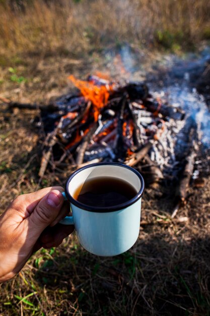 Main tenant une tasse de café à côté d'un feu de camp