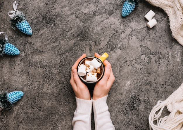 Main tenant une tasse avec des guimauves près de jouets de Noël et une écharpe tricotée