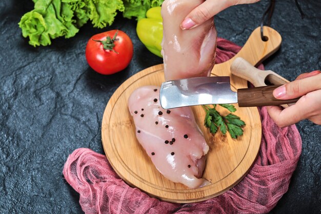 Mains coupant un morceau de filet de poulet cru sur une plaque en bois avec des légumes frais et une nappe.
