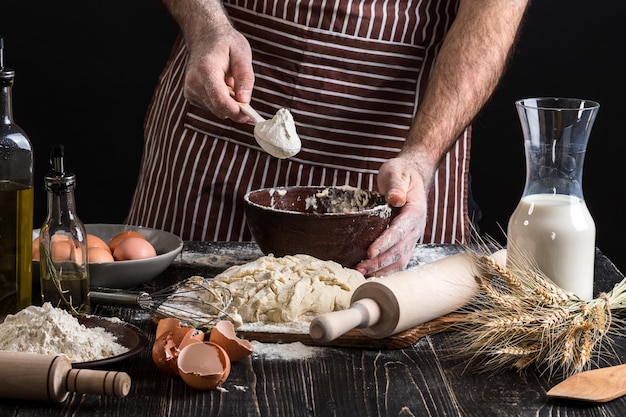 Les mains du chef saupoudrent de la farine dans un bol pour faire de la pâte. Vue rapprochée sur fond noir