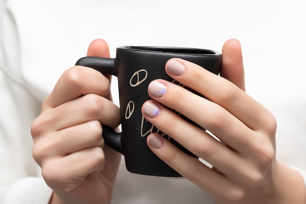 Mains féminines avec un design d'ongle beige tenant une tasse noire.