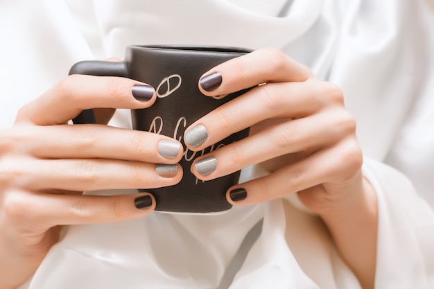 Mains féminines avec des ongles scintillants tenant une tasse noire.