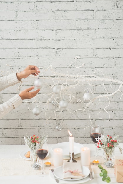 Mains de femme décorer une branche d'arbre avec des boules à côté de la table de Noël arrangée avec goût