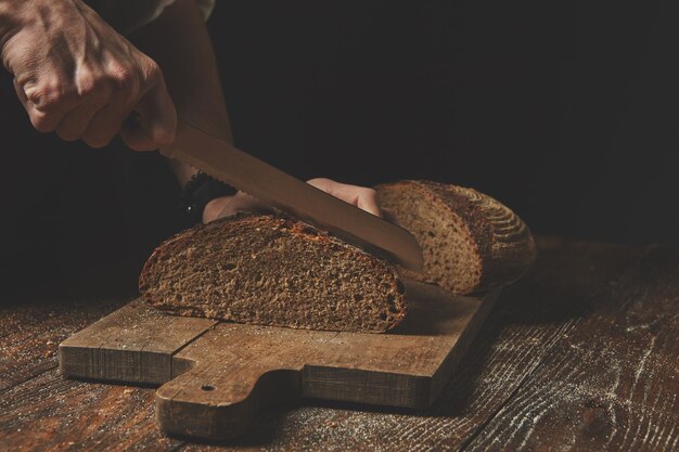 Les mains des hommes coupent maison un pain noir fraîchement cuit sur une planche à découper en bois sur le fond d'une vieille table brune