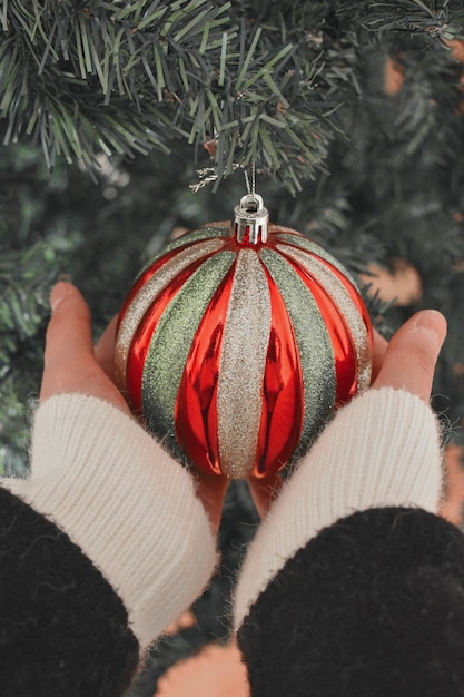 Photo gratuite mains d'une personne tenant une belle boule avec des rayures rouges et dorées sur un arbre de noël