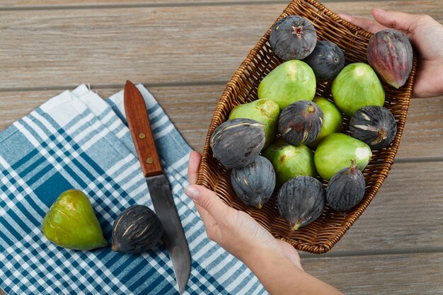 Mains tenant un panier de figues sur table en bois avec un couteau et une nappe.