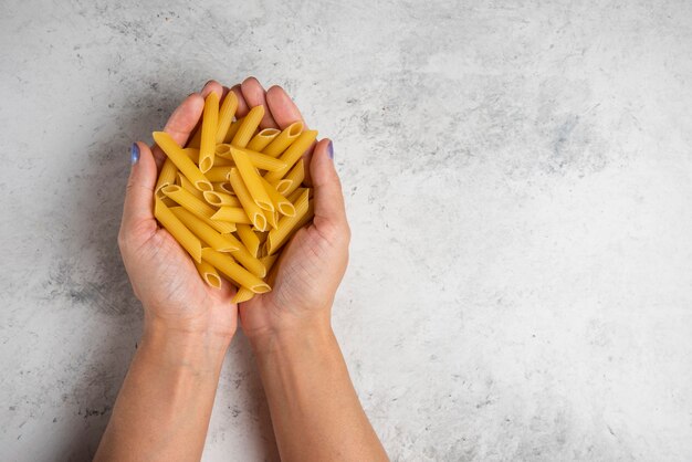 Mains tenant des pâtes penne crues sur fond blanc.