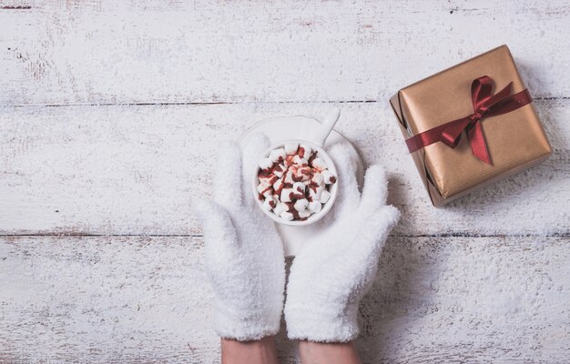 Mains tenant une tasse avec des guimauves avec un cadeau à côté