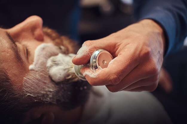 Un maître coiffeur prépare le visage pour le rasage et enduit le visage de mousse dans un salon de coiffure.