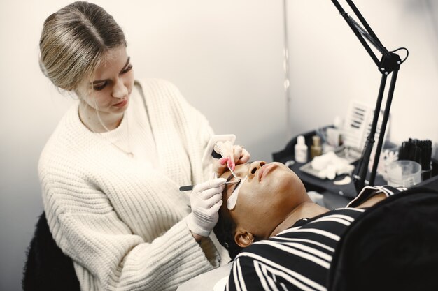 Maître des extensions de cils. Fille africaine faisant la procédure. Femme sur canapé.